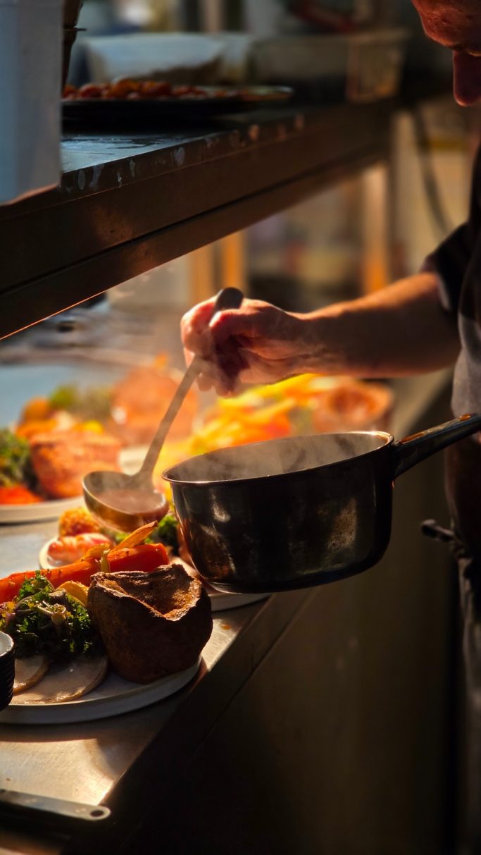 A chef plating a dish with vegetables and sauce in a bustling kitchen.