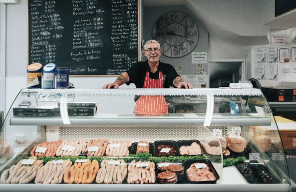 Butcher smiling behind a display of assorted meats and products in a shop.