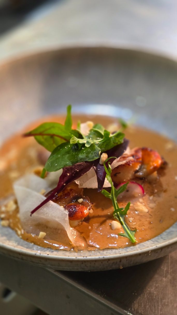 A bowl of spicy soup with vegetables, herbs, and garnishes.